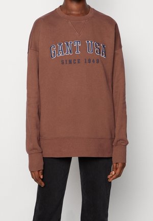 Personne portant un sweat-shirt marron avec "GANT USA Since 1949" en lettres bleu marine et un pantalon noir sur un fond uni.