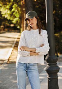 Blusa bianca con tessuto texturizzato e dettagli in pizzo, abbinata a jeans azzurri chiari e un cappellino da baseball verde scuro. In piedi contro un palo.