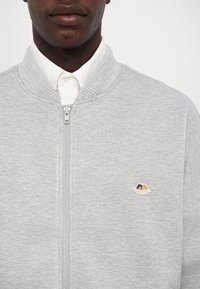 Grijze jas met ritssluiting, geribde kraag en manchetten, met een klein geborduurd ontwerp op de linkerborst. Gemaakt van zachte, gestructureerde stof.