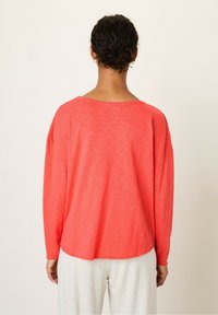 Langærmet, koralfarvet pullover med en afslappet pasform. Blød tekstur, bred udskæring og let buet kant. Matchet med lysegrå bukser.