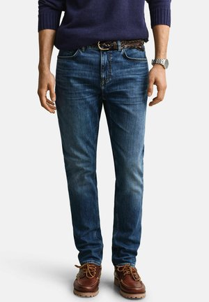 Man draagt een donkerblauwe trui, nauwsluitende blauwe spijkerbroek met een gevlochten bruine riem, bruine leren schoenen en een zilveren polshorloge aan de linkerpols.