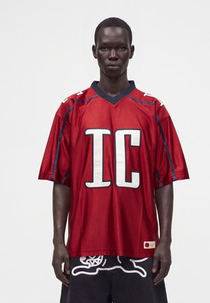 Hombre de piel oscura con una camiseta deportiva roja holgada con grandes letras blancas "IC" y pantalones cortos negros con diseño blanco, de pie frente a un fondo liso.