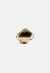 Vaalean beige-koruton kenkä, jossa on pyöreä nenä, koristeltu strasseilla etuosassa ja sivuilla. Kengässä on matala korko ja sileä pinta.