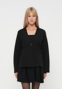 Blond kvinna iklädd en svart blazer med en knapp över en svart plisserad kjol och svarta strumpbyxor, stående mot en enfärgad vit bakgrund.