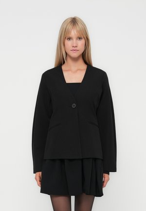 Blonde woman wearing a black single-button blazer over a black pleated skirt and black tights, standing against a plain white background.