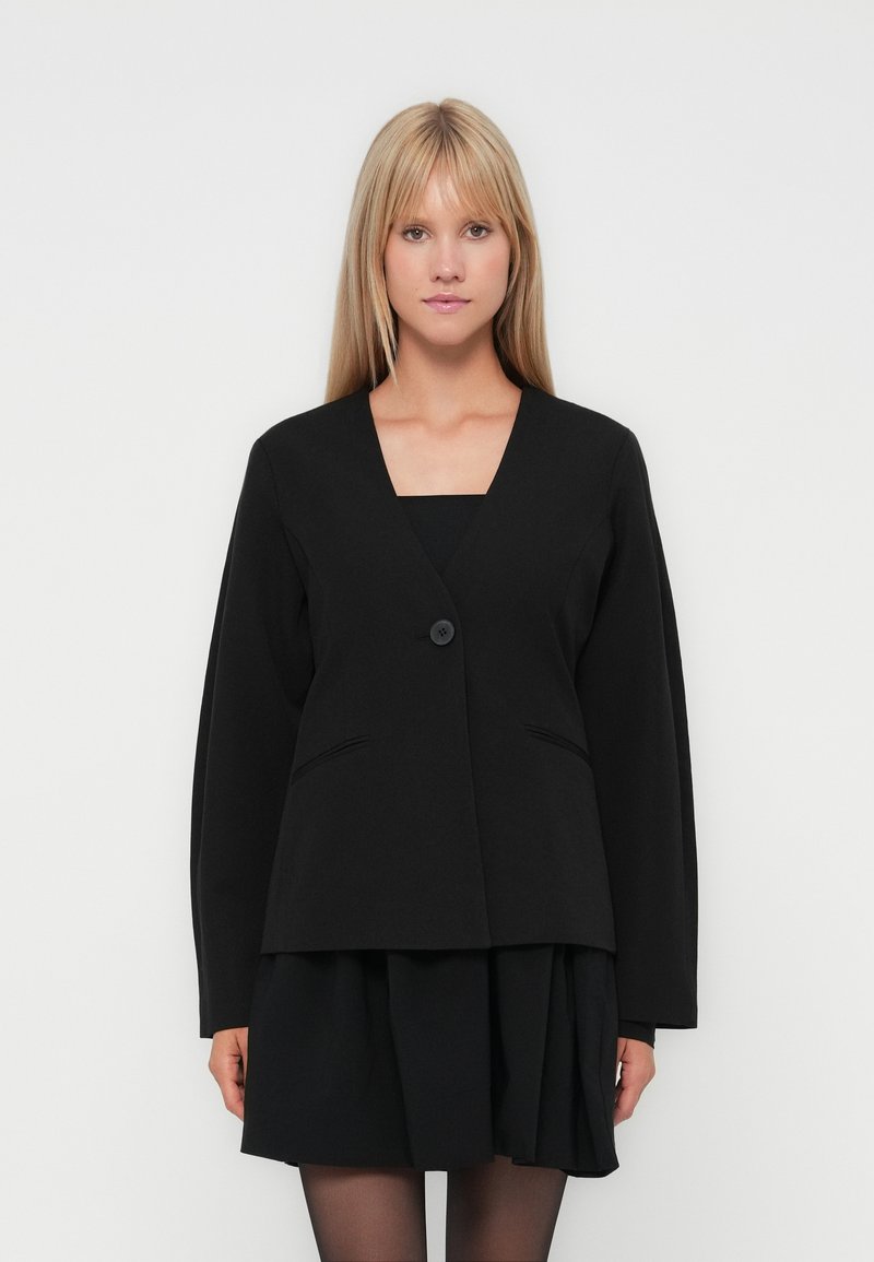 Blond kvinna iklädd en svart blazer med en knapp över en svart plisserad kjol och svarta strumpbyxor, stående mot en enfärgad vit bakgrund.