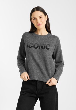 Grijze gebreide trui met het woord "ICONIC" in het zwart, lange mouwen, geribbelde boorden en een licht cropped zoom. Draagt het met zwarte jeans.