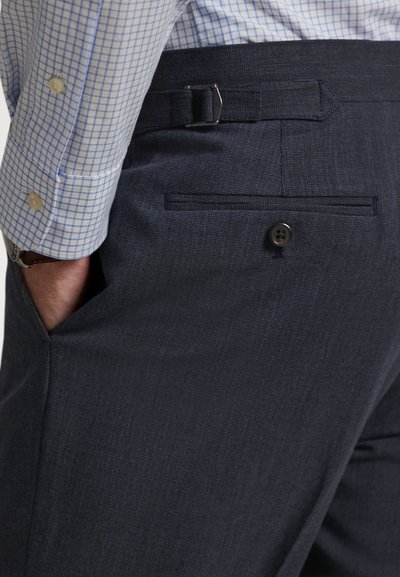 Vyras dėvi tamsiai mėlynas kelnes su šoniniu reguliatoriumi ir nugaros kišenėle su sagomis, ranka įkišta į šoninę kišenę, šviesiai mėlyną languotą marškinių modelį.