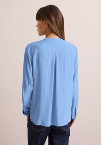 Blå långärmad blus i lätt tyg med rak skärning, veckdetalj bak och rundad nederkant. Bärs med mörka jeans.