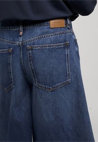 Vue de dos d'une personne portant un jean en denim bleu foncé ample avec des coutures visibles, des poches arrière et une étiquette en cuir marron sur la ceinture.