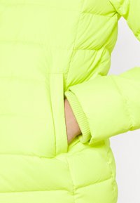 Doudoune jaune néon avec une poche, des poignets côtelés et une texture matelassée avec des coutures horizontales pour l'isolation.