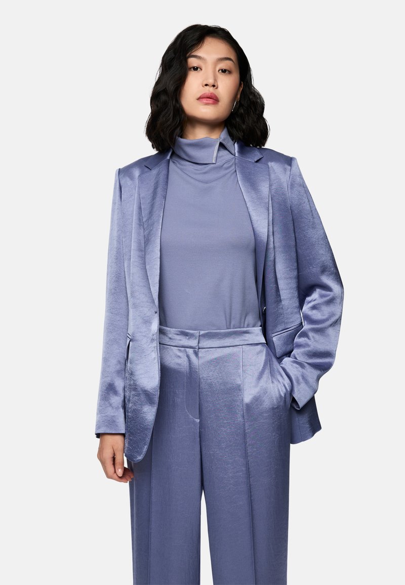 Woman with shoulder-length dark hair wearing a shiny lavender blazer, matching wide-leg pants, and a high-neck top, standing with one hand in pocket.