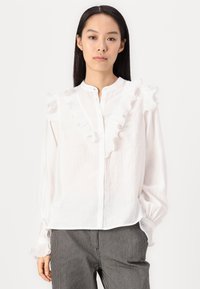 Hvit bluse med volangdetaljer langs fronten og skuldrene. Har knappelukking og lange ermer. Materialet virker lett og teksturert.