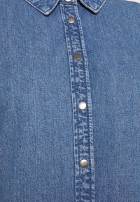 Chemise en denim bleu avec un col classique, des boutons en métal et une texture subtile. Le tissu présente un tissage homogène avec un dégradé minimal.