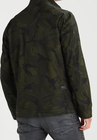 Man med mörkgrön och svart kamouflagejacka i kombination med urtvättade svarta jeans, sedd bakifrån från midjan och uppåt.