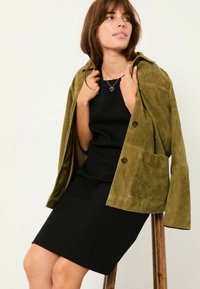 Olivegrüne Wildlederjacke mit Kragen, Fronttaschen und Knopfschluss, über einem enganliegenden schwarzen Kleid getragen. Schlichtes und texturiertes Design.