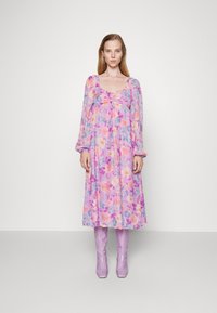 Vestido maxi floral em tecido leve, com uma base roxa e flores em tons pastel, detalhe de pregas no peito e mangas longas. Combinado com botas lilás até ao joelho.