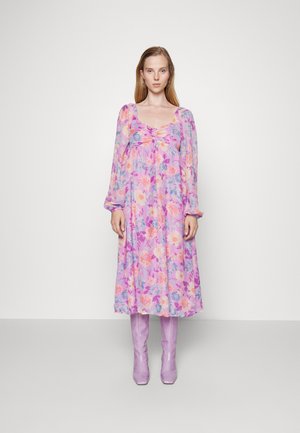 Vestido maxi floral em tecido leve, com uma base roxa e flores em tons pastel, detalhe de pregas no peito e mangas longas. Combinado com botas lilás até ao joelho.