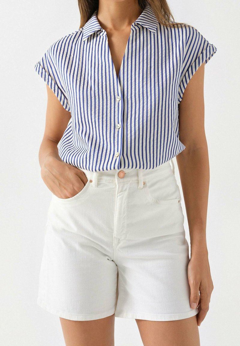 Hemd mit Knöpfen in Creme mit blauen vertikalen Streifen, kurzen umgeschlagenen Ärmeln, kombiniert mit weißen Denim-Shorts mit einer Fronttasche.