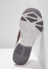 Suola di una scarpa sportiva marrone e bianca con motivi di battistrada geometrici, inclinata contro un angolo di un muro e del pavimento bianchi.