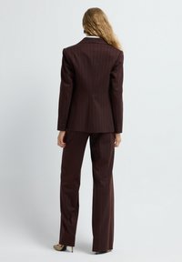 Mujer con cabello largo y rubio, vestida con un traje a rayas marrón oscuro y tacones beige, de pie de espaldas contra un fondo claro y liso.