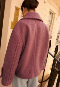 Veste en polaire violette surdimensionnée avec une surface texturée, un large col et des poches latérales, associée à un pantalon bleu clair.