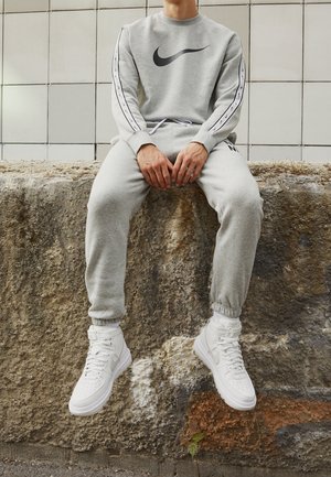 Osoba w szarym dresie Nike i białych wysokich butach sportowych Nike siedzi na szorstkiej kamiennej krawędzi przy ścianie wyłożonej kafelkami.