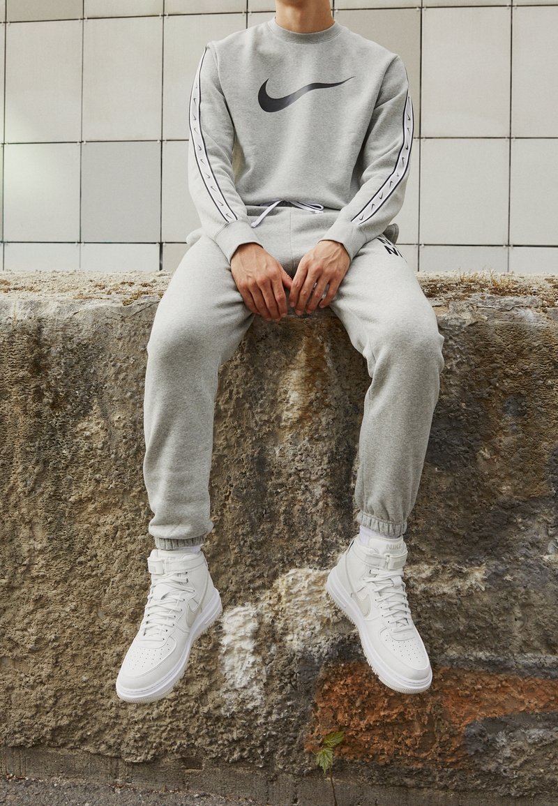 Personne portant un survêtement gris Nike et des baskets montantes blanches Nike assise sur un rebord en pierre rugueuse contre un mur carrelé.