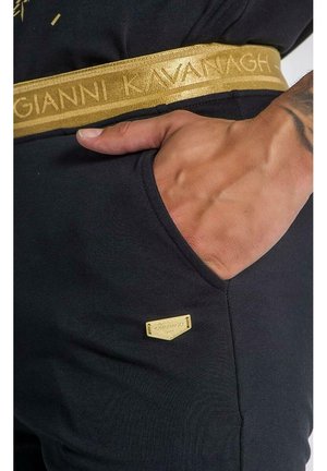 Main reposant dans la poche d’un pantalon noir avec une ceinture dorée et une petite étiquette de marque dorée portant l’inscription « Kavanagh ».