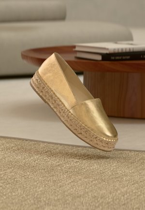 Espadrille dorée à enfiler avec semelle plateforme en jute tressé, présentée sur un tapis beige devant une table basse en bois avec des livres empilés.