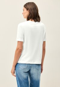 T-shirt bianco con maniche corte, dal fit rilassato e colletto rotondo. Il tessuto ha una texture liscia, mostrato da dietro con jeans blu.