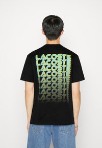 Black cotton T-shirt featuring a large, multicolored "LACOSTE" graphic printed vertically on the back, with a textured finish.