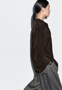 Mørkebrun satinbluse med afslappet pasform, detaljer med sideplissering og knapgående stykke, parret med lysegrå vidde bukser.