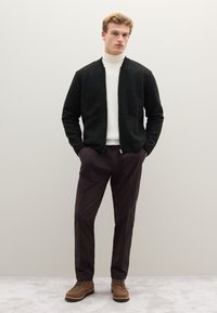 Cardigan nero lavorato a maglia con due tasche frontali, dolcevita bianco, pantaloni marroni e stivali marroni con lacci. Tessuto liscio, design minimale.