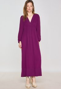 Vestito lungo di un profondo colore viola con scollatura a V, maniche lunghe e vita arricciata, caratterizzato da un tessuto morbido e un orlo dritto. Indossato con espadrillas.