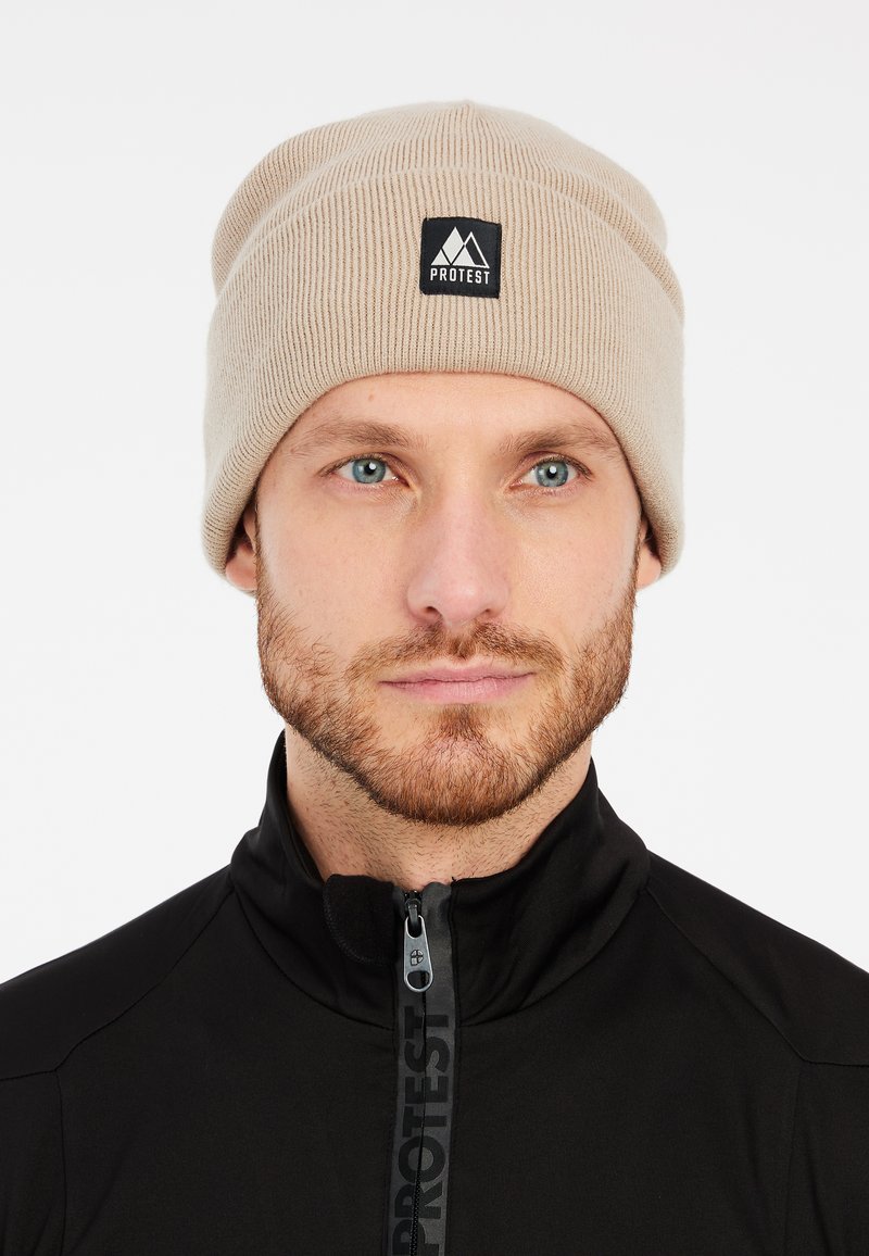 Beige knitted beanie with ribbed texture, featuring a black square patch with the logo "PROTEST." Worn with a zippered black collar jacket.