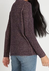 Violetti neulepaita pitkillä hihoilla, jossa on teksturoitu muotoilu. Helma on hieman pidempi takana, ja se on yhdistetty vaaleansinisiin farkkuihin.