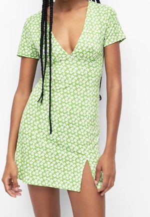 Femme portant une robe verte à manches courtes avec de petites fleurs blanches et une fente avant, se tenant devant un fond blanc uni.