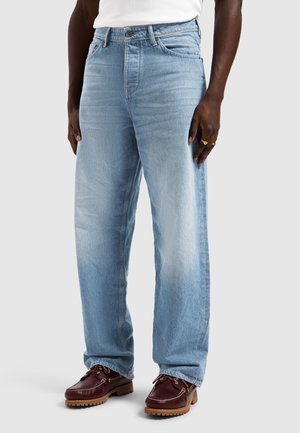 Man draagt lichtblauwe jeans met wijde pijpen, wit overhemd, bruine leren casual schoenen en gouden ringen aan de rechterhand, tegen een effen achtergrond.