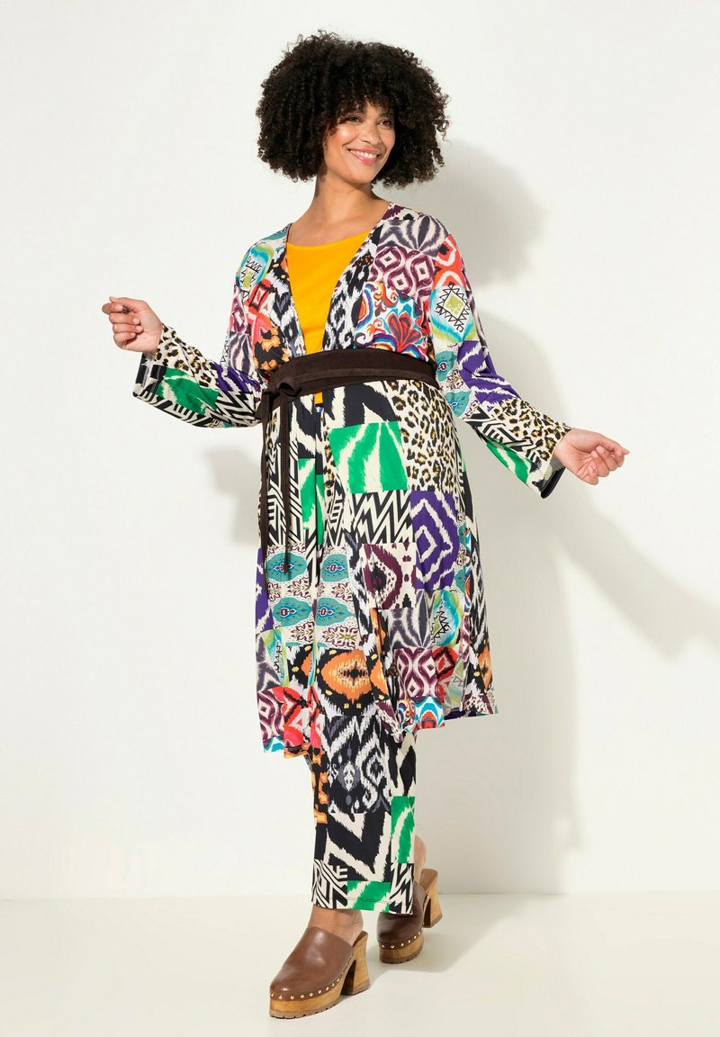 Giacca kimono a maniche lunghe con fantasia colorata, cintura marrone, abbinata a una maglietta gialla e pantaloni fantasia, caratterizzati da motivi geometrici e animali diversi.