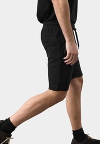 Man walking wearing black drawstring shorts, black socks, and dark shoes against a plain light background.