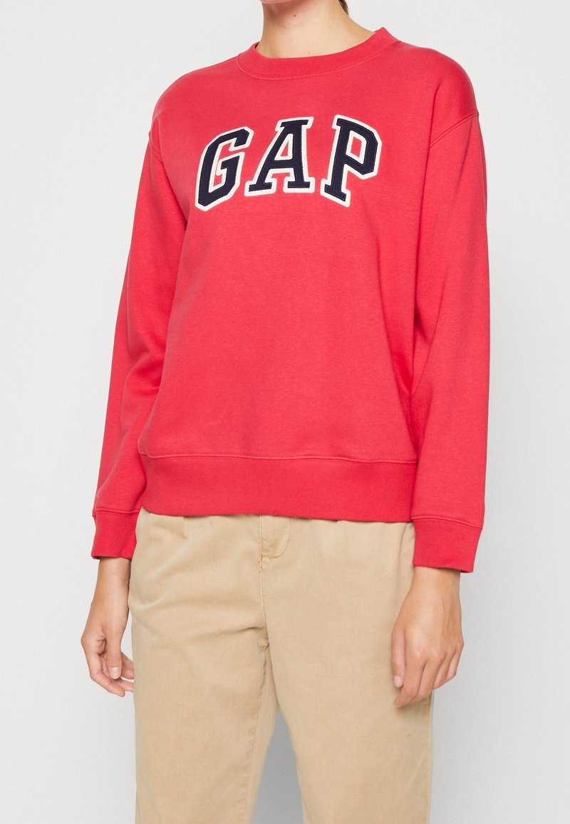 Femme portant un sweat rouge GAP avec des lettres bleu marine et blanches et un pantalon beige, debout devant un fond clair uni.