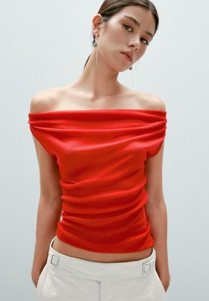 Off-the-shoulder red top with a fitted, ruched design. Smooth fabric and glossy texture, paired with white pants featuring a belt accent.