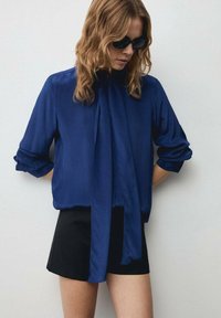 Blusa di seta blu navy con cravatta al collo, maniche lunghe arrotolate fino ai gomiti, indossata con pantaloni corti neri. Texture liscia, design minimalista.