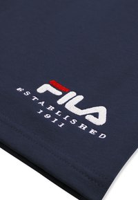 Zbliżenie na granatową tkaninę z wyhaftowanym białym i czerwonym logo FILA oraz napisem "Established 1911".