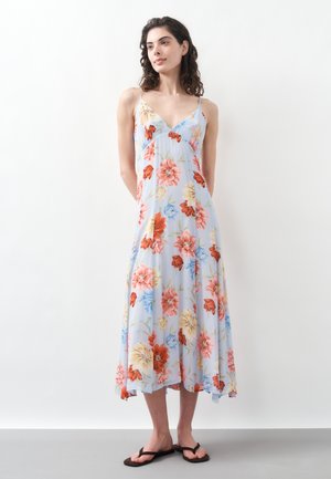 Femme debout portant une robe midi sans manches bleu clair avec un imprimé floral rouge, jaune et bleu, et des sandales tong noires.