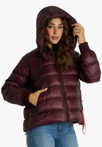 Donna che indossa una giacca a vento bordeaux lucido con il cappuccio alzato, una mano regge il cappuccio mentre l'altra è in tasca, abbinata a jeans blu.