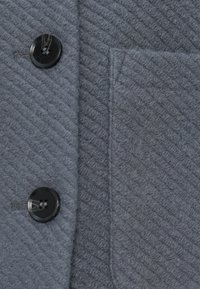 Tessuto di cappotto grigio in lana con due bottoni neri e una tasca verticale cucita sul lato destro.