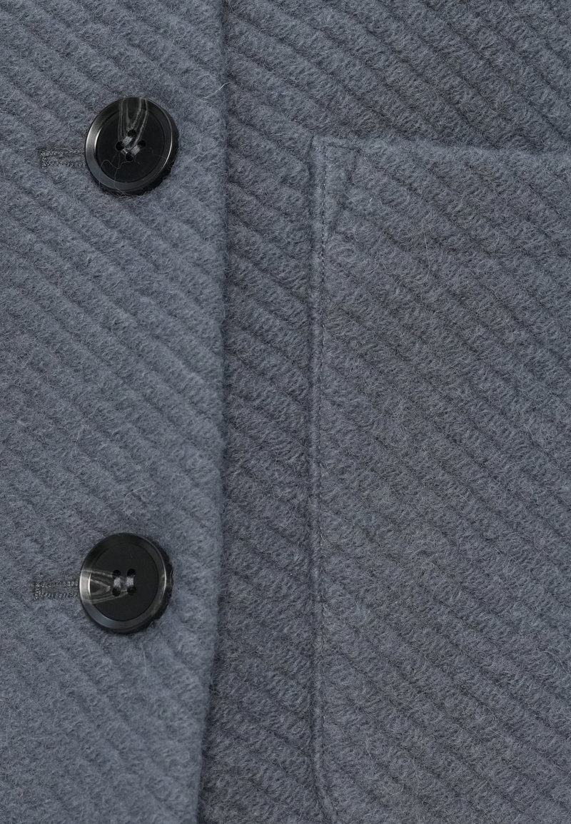 Grauer Wollstoffmantel mit zwei schwarzen Knöpfen und einer genähten senkrechten Tasche auf der rechten Seite.