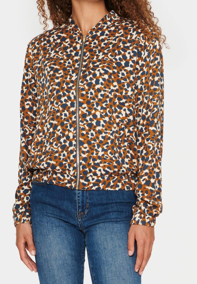 Kvinna som bär en zip-up jacka med ett brunt, marinblått och vitt abstrakt leopardmönster, matchat med blå jeans.